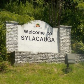 A welcoming 'Welcome to Sylacauga' sign board surrounded by lush, green trees